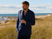Man in a navy suit standing in a grassy field with ocean and sky in the background