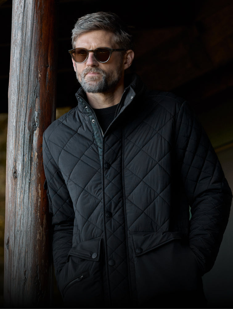 Man wearing a black quilted jacket and sunglasses against a dark background