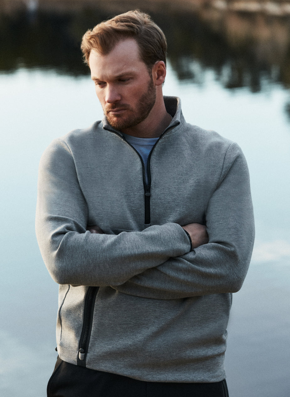 Man wearing a gray quarter-zip hoodie standing by a body of water